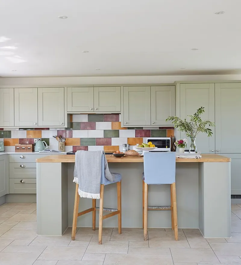 Bright modern kitchen with sage-green cabinets, colourful tiled splashback and central island with stools, overlooking open countryside