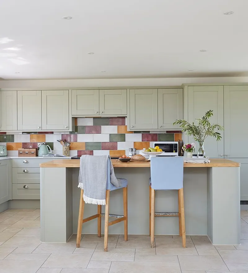 Bright modern kitchen with sage-green cabinets, colourful tiled splashback and central island with stools, overlooking open countryside