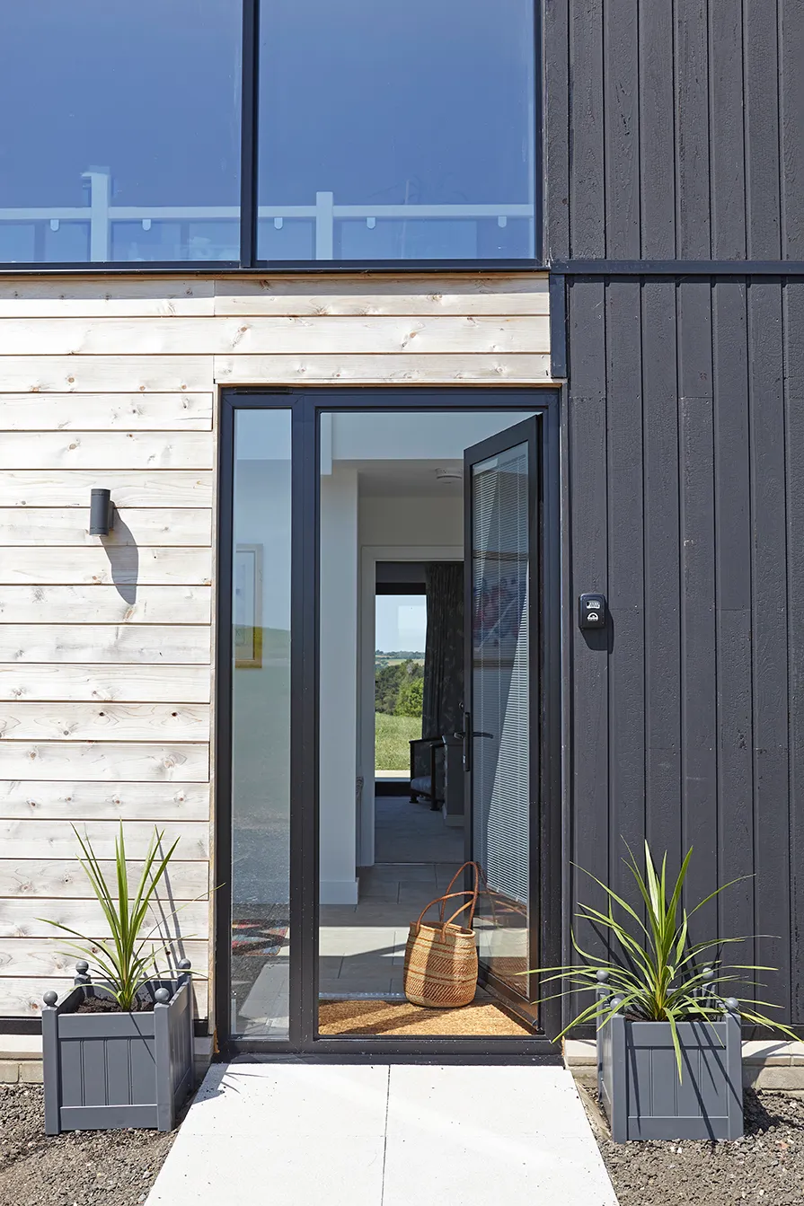 Modern timber-clad entrance with open glass door, planters and woven basket on doormat