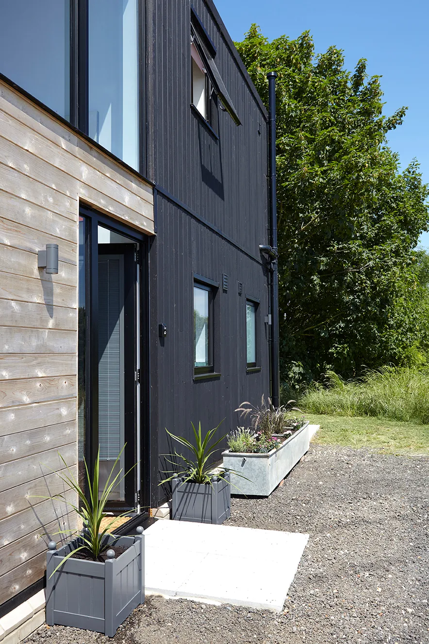 Modern house exterior with black cladding and timber wall beside a glazed entrance, planters and a gravel path