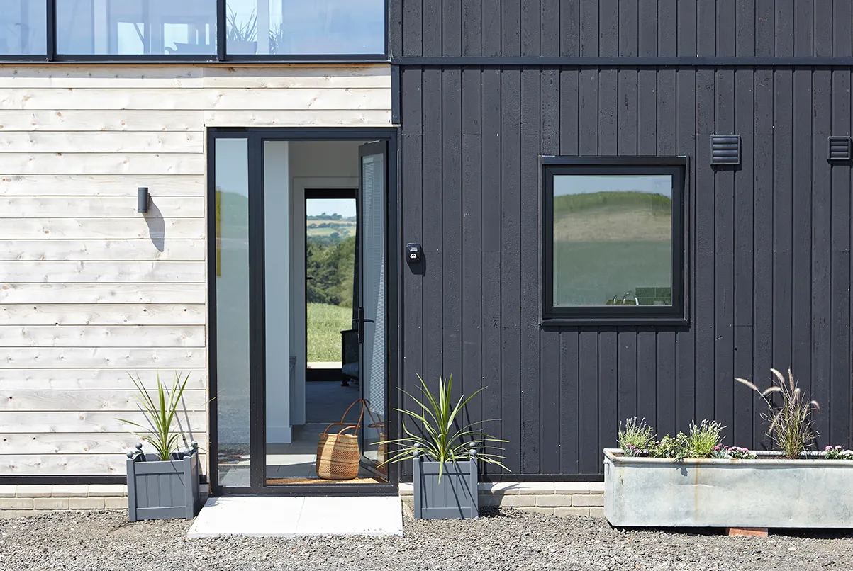 Modern timber-clad house entrance with open glass door, black window frame and planters