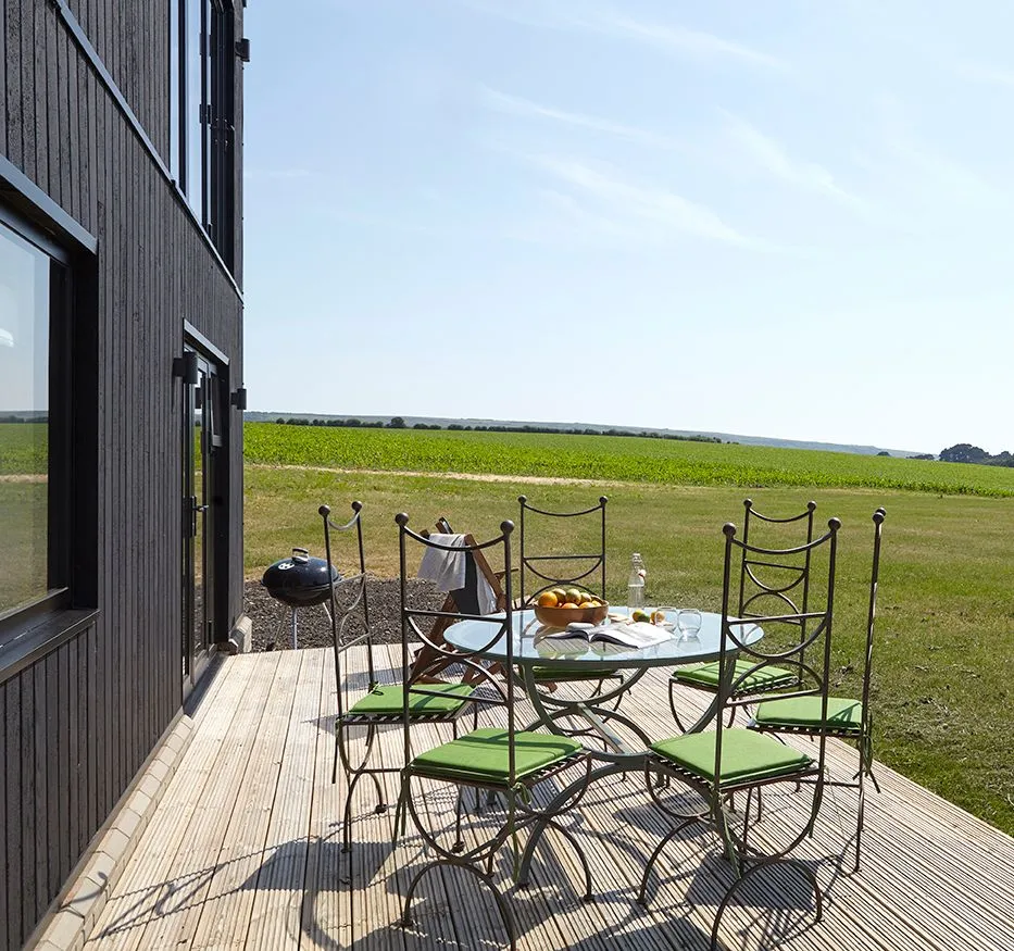 Outdoor dining table with green-cushioned chairs on a wooden deck beside a modern house, overlooking open countryside