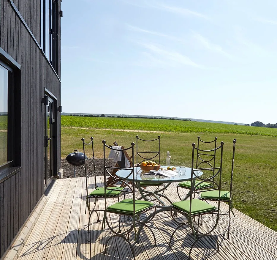 Outdoor dining table with green-cushioned chairs on a wooden deck beside a modern house, overlooking open countryside