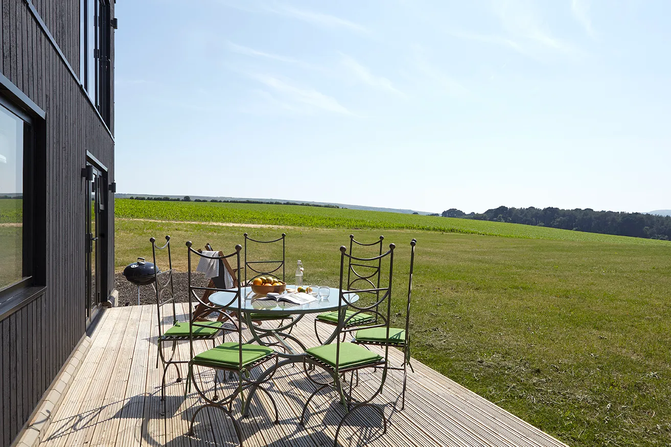 Outdoor dining table with green-cushioned chairs on a wooden deck beside a modern house, overlooking open countryside