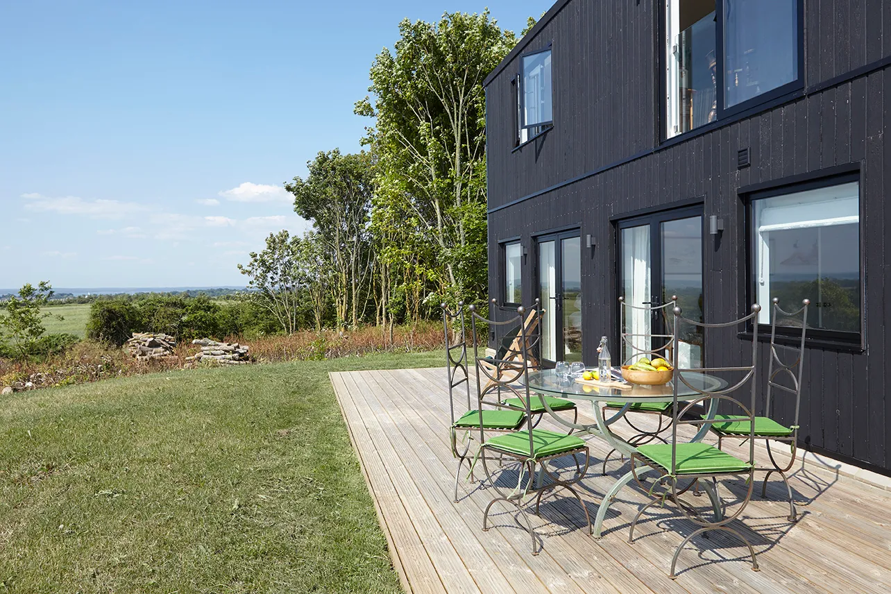 Modern black-clad house with a wooden terrace and outdoor dining table overlooking countryside views