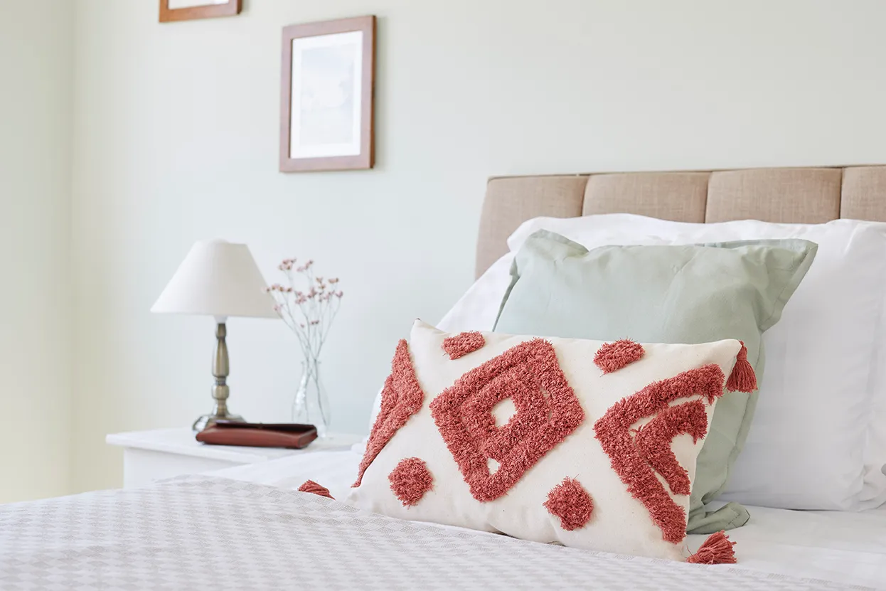 Cosy bedroom with neutral bedding, sage green pillows and a patterned cushion, beside a bedside table with lamp and flowers