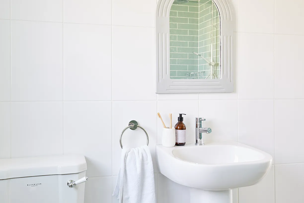 Bright white tiled bathroom with pedestal sink, chrome tap, towel ring and wall mirror