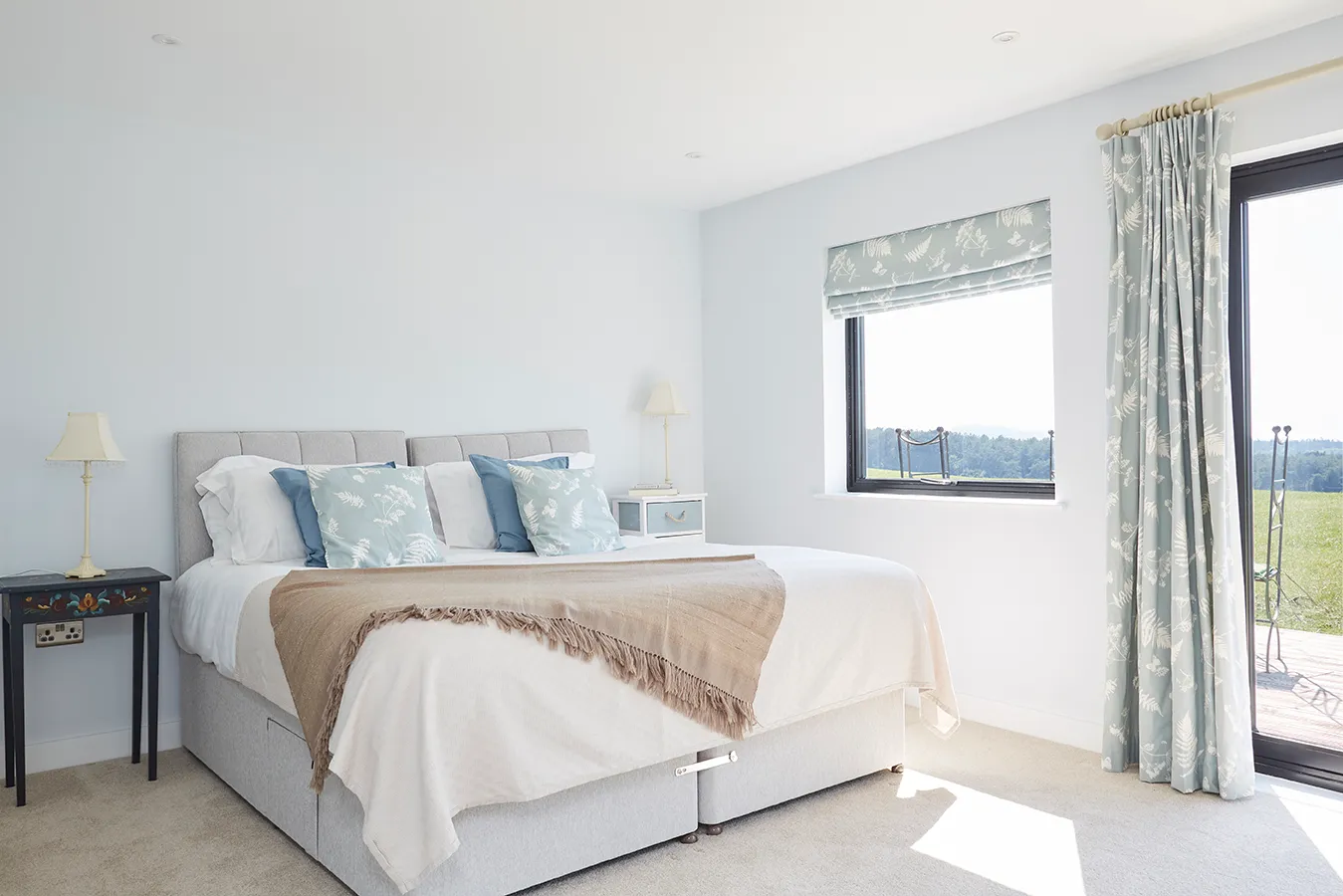 Bright, airy bedroom with double bed, blue patterned cushions and curtains, and large windows with countryside views