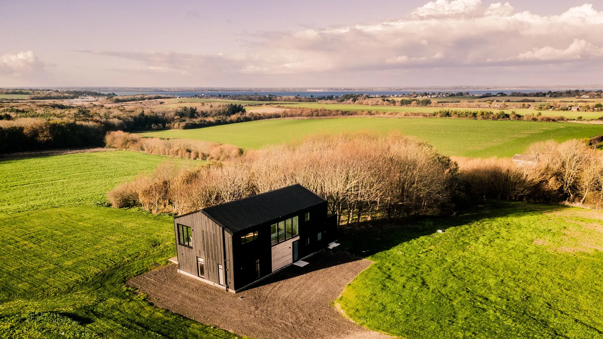 Aerial view of a modern black-clad wendy house on a hillside surrounded by green fields and winter trees