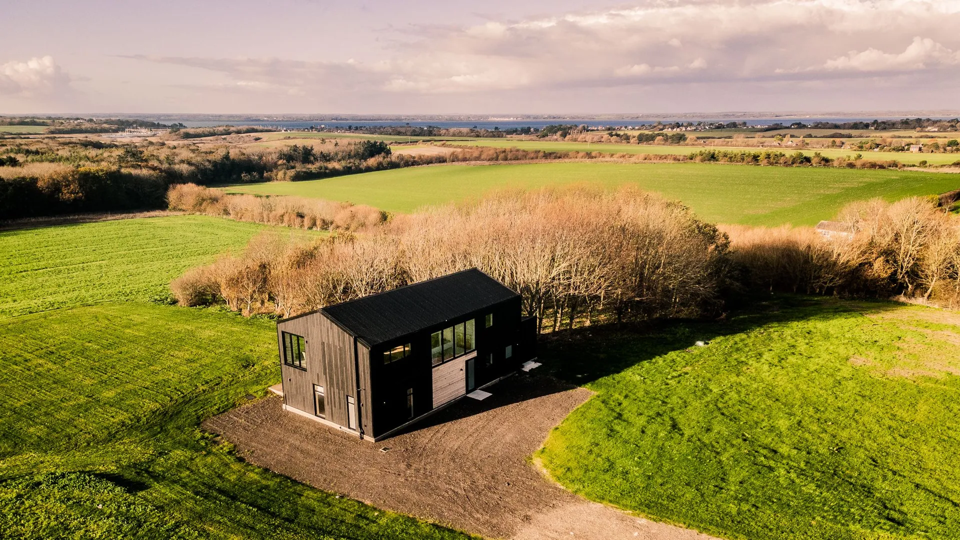 Aerial view of a modern black-clad wendy house on a hillside surrounded by green fields and winter trees