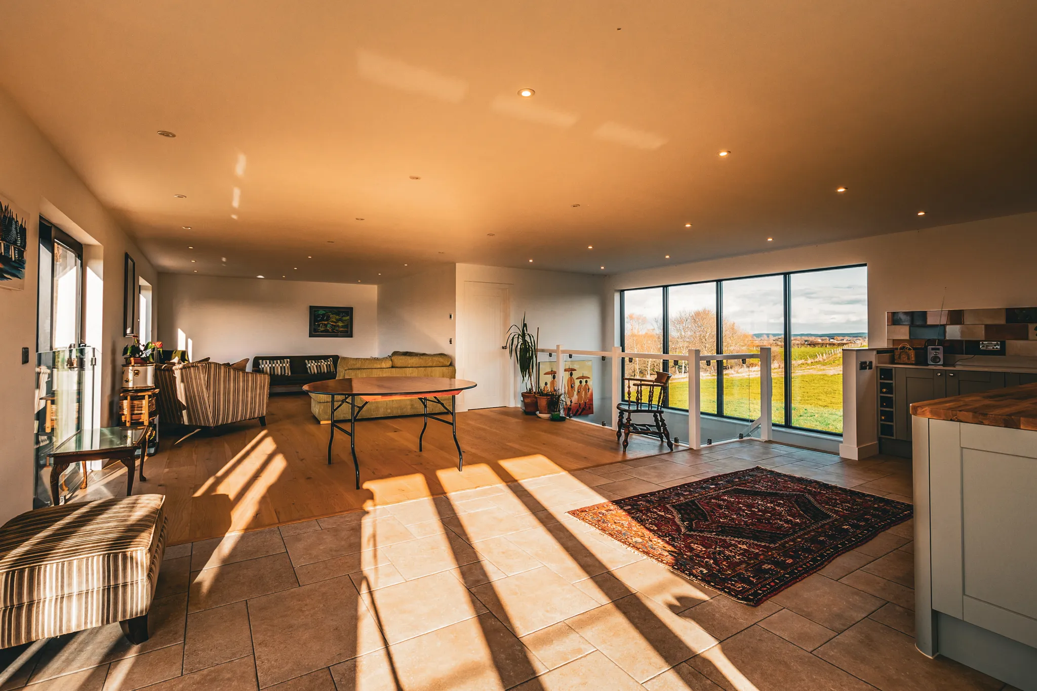 Sunlit open-plan living area with sofa seating, wooden table and large windows overlooking countryside