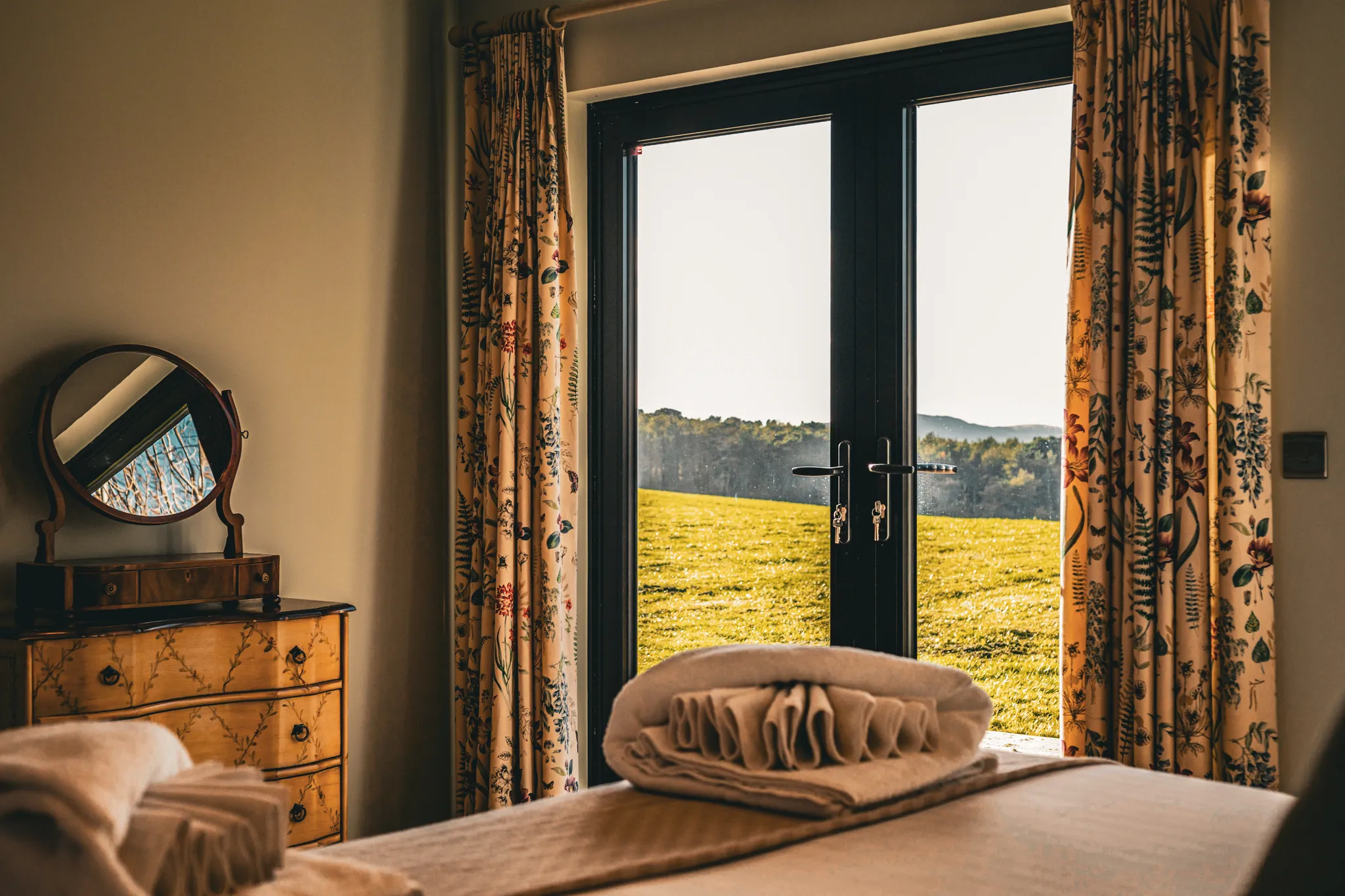 Cosy bedroom with folded towels on the bed and French doors opening onto a sunlit countryside view