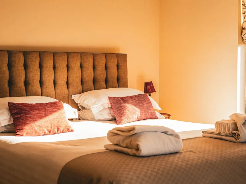 Cosy bedroom with double bed, tufted headboard, red cushions and folded towels in warm sunlight