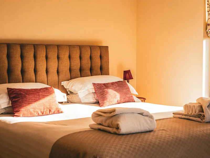 Cosy bedroom with double bed, tufted headboard, red cushions and folded towels in warm sunlight