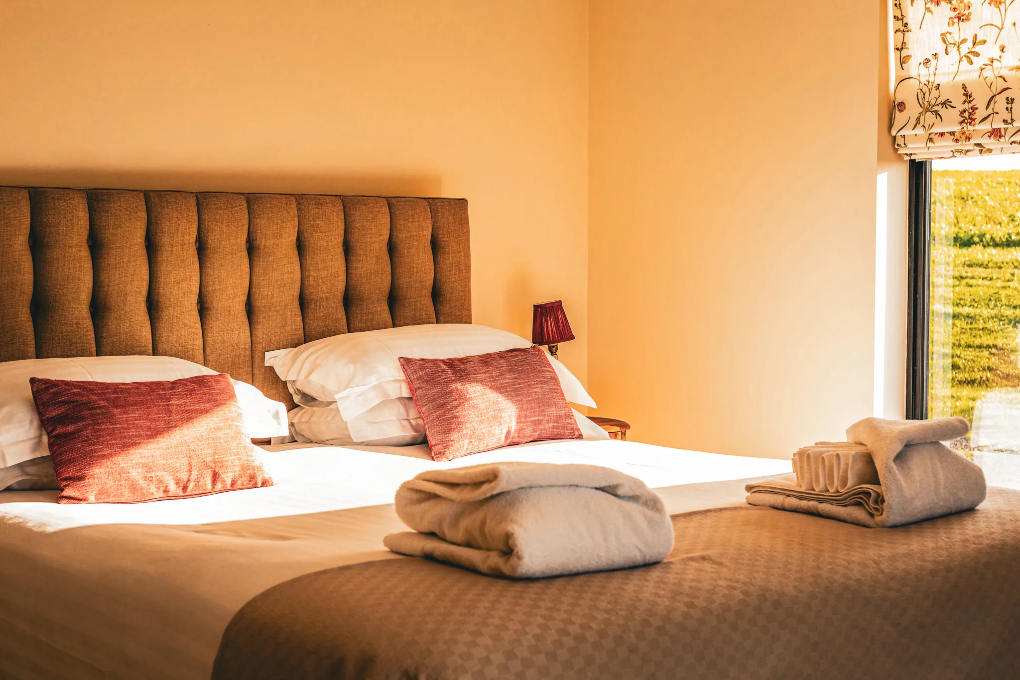 Cosy bedroom with double bed, tufted headboard, red cushions and folded towels in warm sunlight