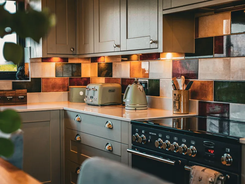 Cosy kitchen corner with grey cabinets, colourful tiled splashback, and sage green toaster and kettle beside a black range cooker