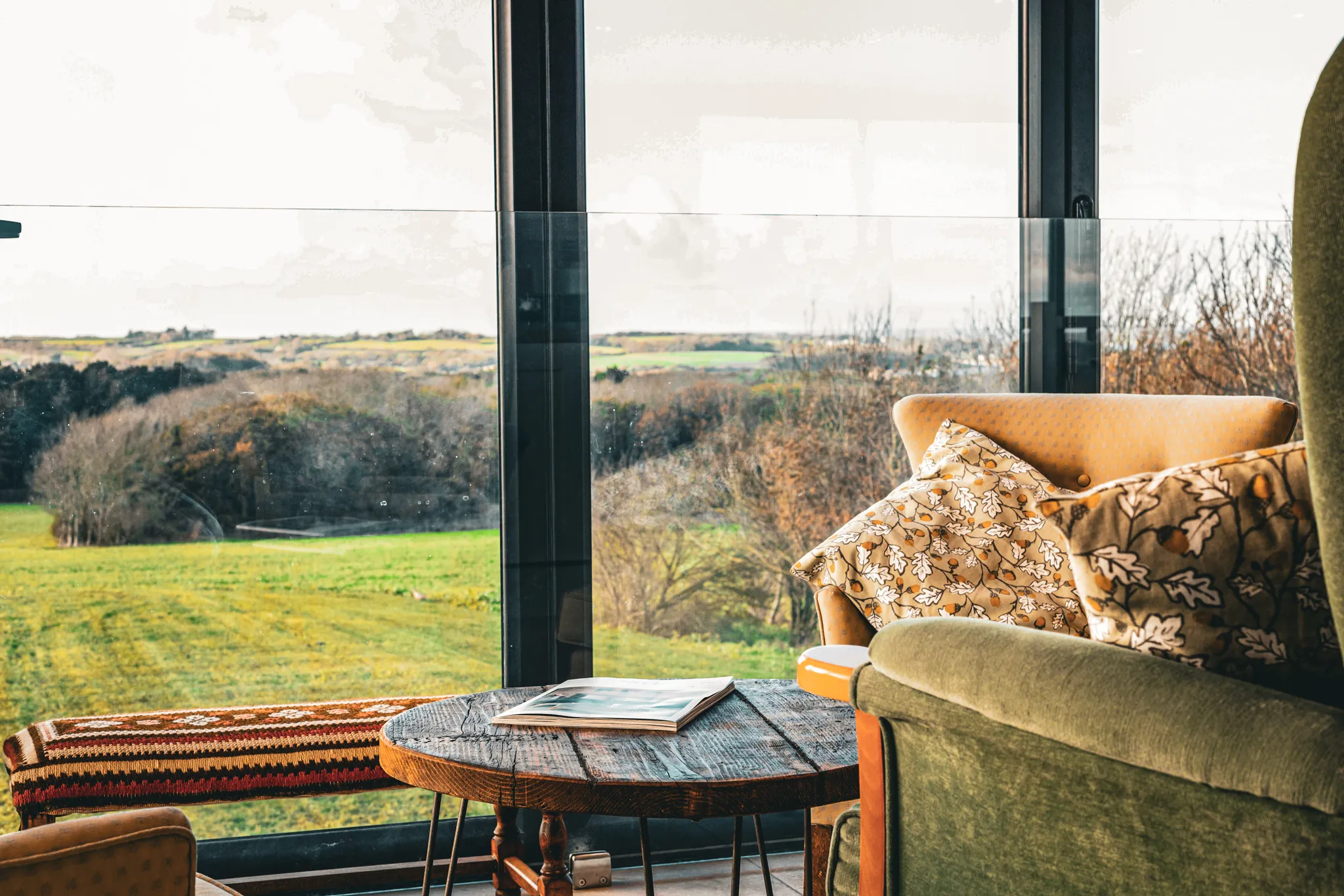 Cosy seating area with armchairs and patterned cushions beside a rustic coffee table, looking out through large windows over rolling countryside