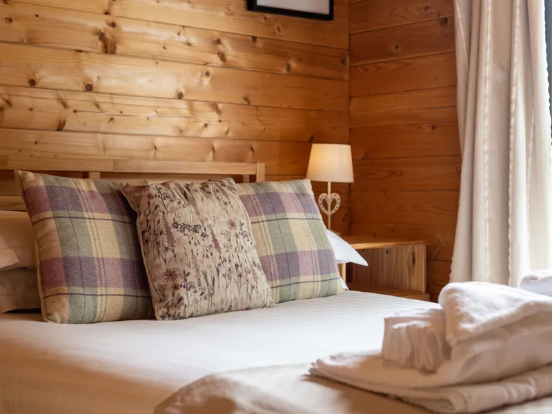 Cosy wooden cabin bedroom with neatly made bed, plaid and floral cushions, bedside lamp and folded towels