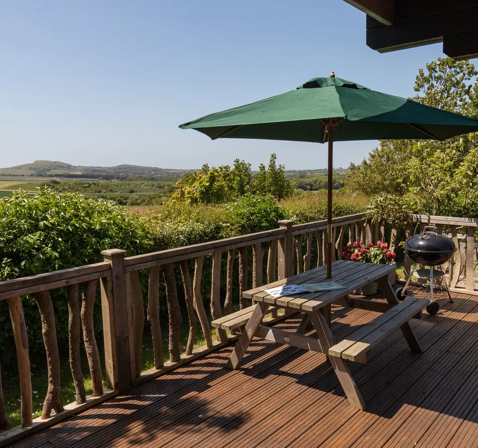 Wooden cabin deck with picnic table, green parasol and barbecue overlooking countryside