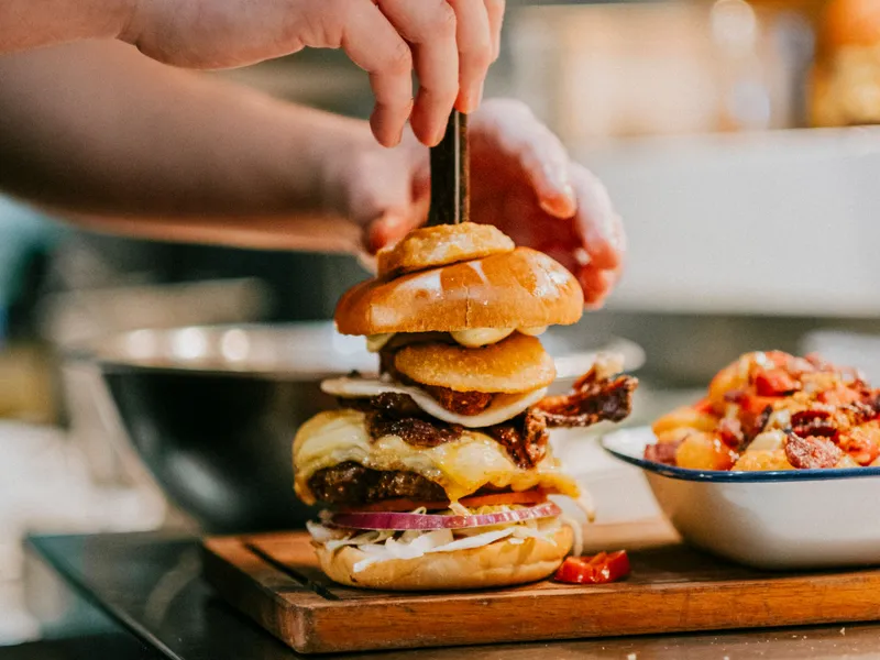 Hands assembling a tall cheeseburger with onions and bacon on a wooden board in a restaurant kitchen