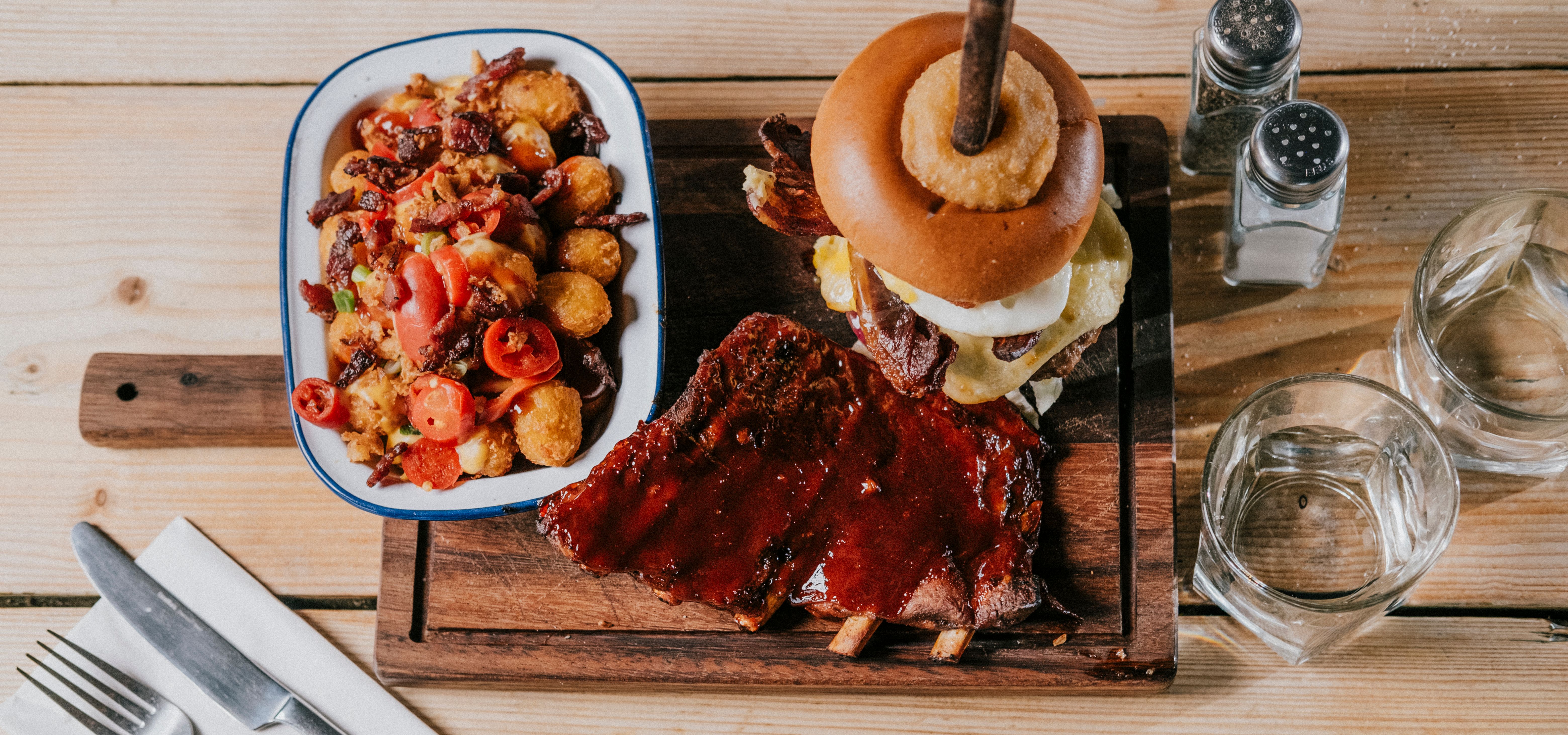 Barbecue ribs and a stacked cheeseburger with onion ring on a wooden board, with loaded tater tots and water on a wooden table