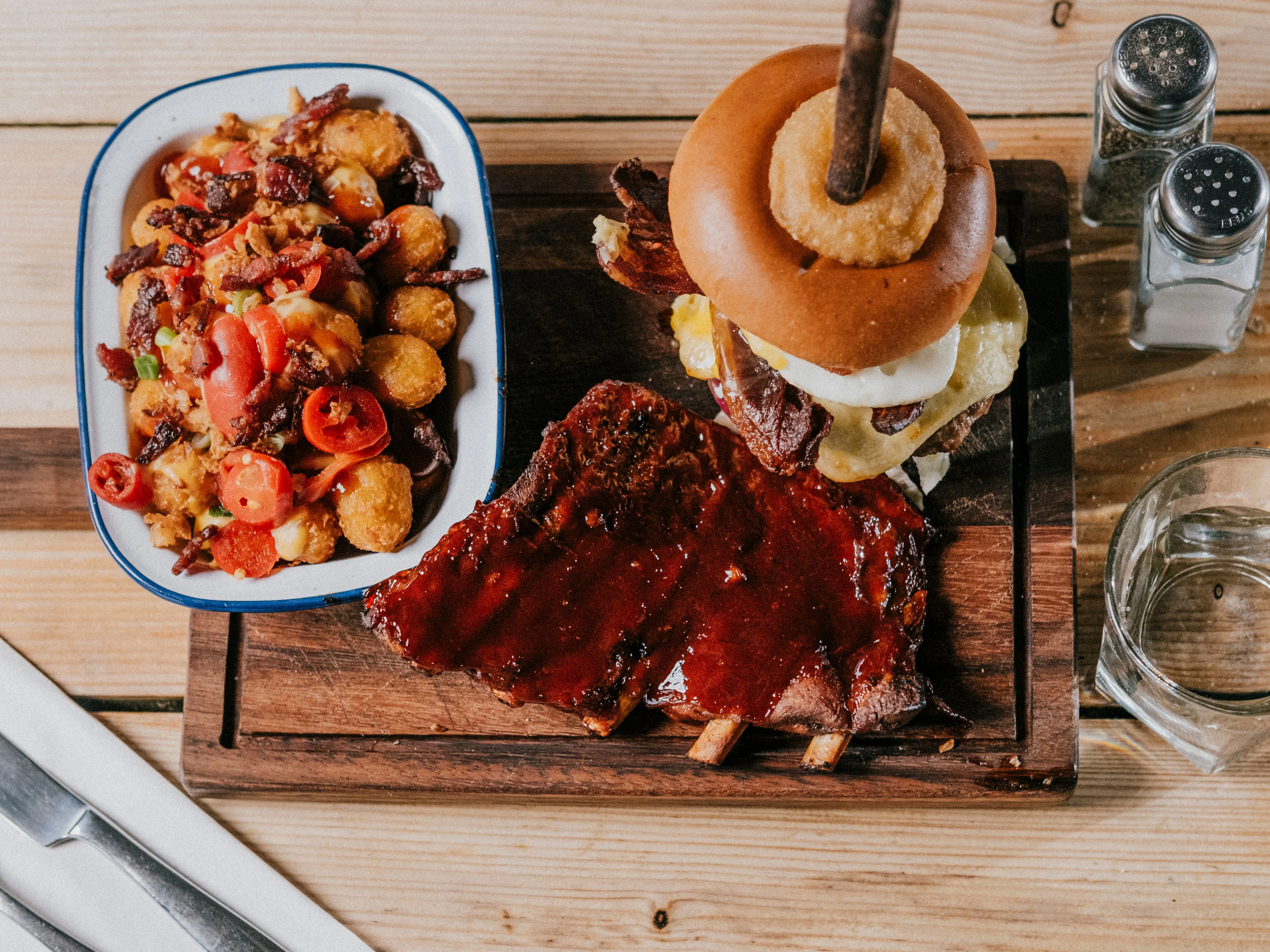 Barbecue ribs and a stacked cheeseburger with onion ring on a wooden board, with loaded tater tots and water on a wooden table