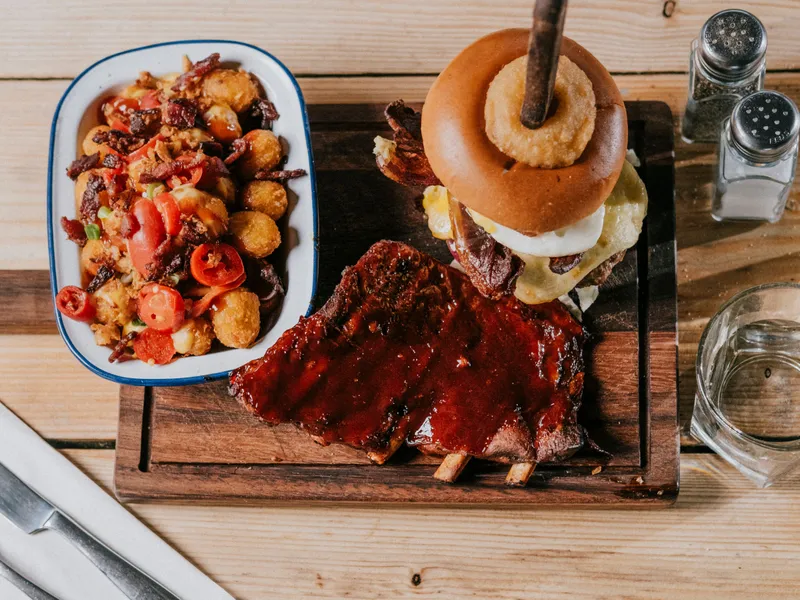 Barbecue ribs and a stacked cheeseburger with onion ring on a wooden board, with loaded tater tots and water on a wooden table