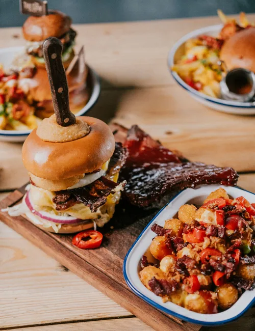 Gourmet burger with crispy onion ring and knife skewer, served with sauced ribs and loaded tater tots on a wooden board