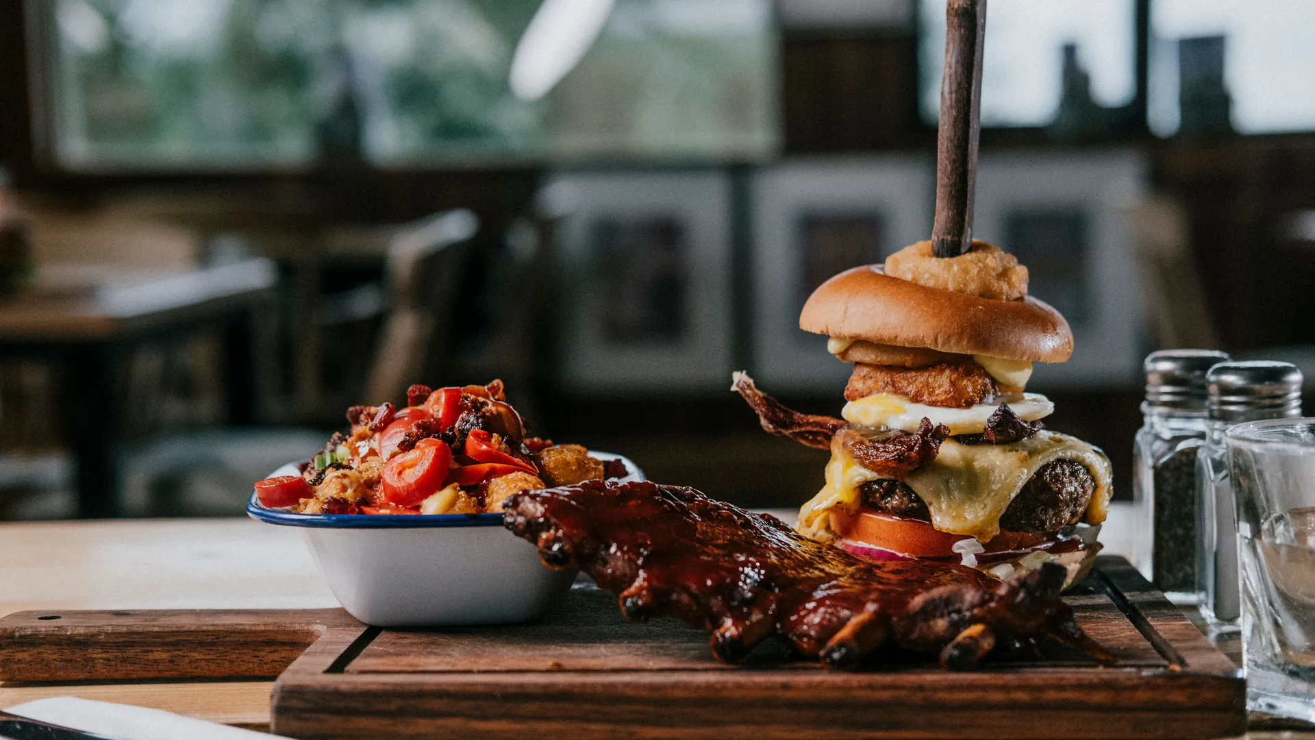 Stacked beef burger with melted cheese, bacon and onion ring, served with barbecue ribs and a salad on a wooden board