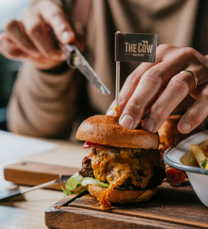 Hand holding a cheeseburger on a wooden board with a small flag reading 