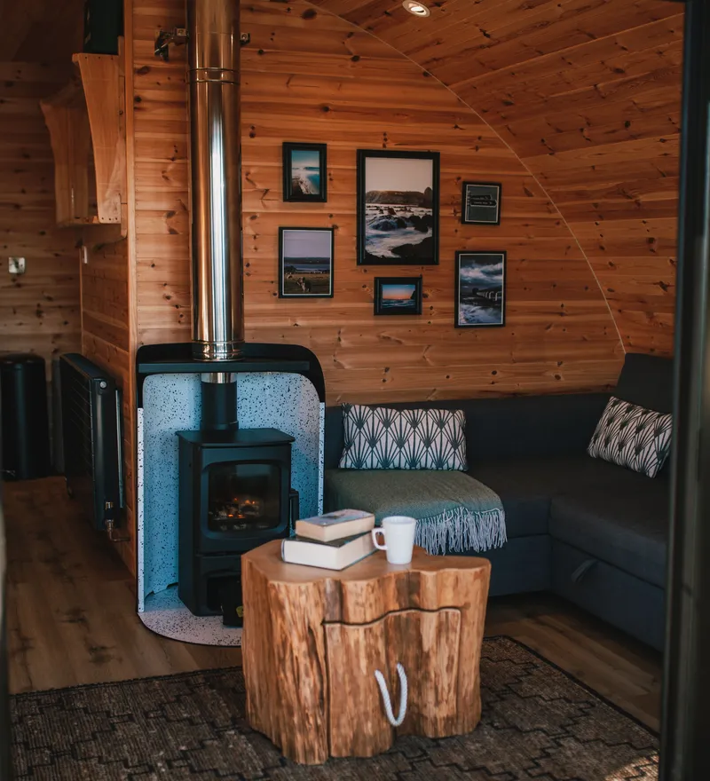 Cozy wooden cabin interior with wood stove, sectional sofa, rustic table, and framed landscape photos on the wall.
