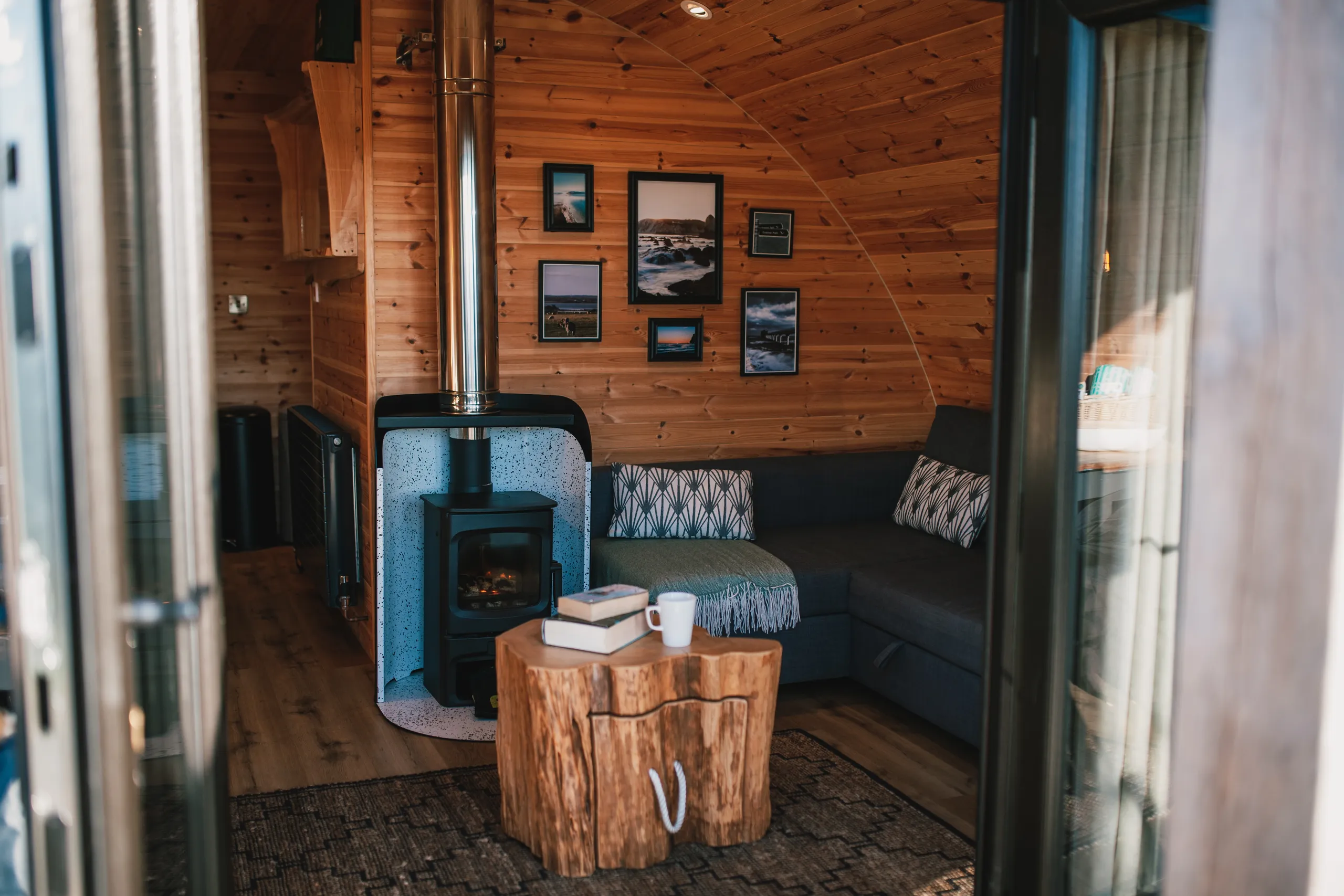 Cozy wooden cabin interior with wood stove, sectional sofa, rustic table, and framed landscape photos on the wall.