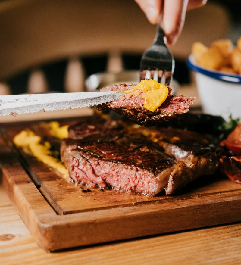 Hand cutting a slice of medium-rare sirloin steak topped with mustard on a wooden board