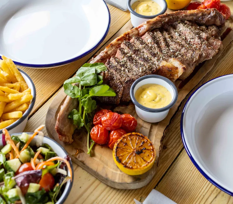 Grilled tomahawk steak on a wooden board with chips, salad, roasted tomatoes and peppercorn sauce