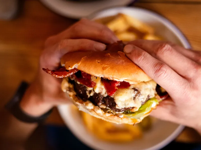 Hands holding a beef burger with melted cheese and bacon over a plate of chips