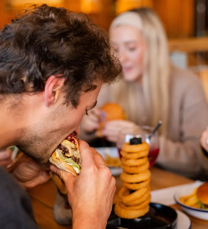 Man eating a burger at a wooden table with onion rings and two women dining in the background