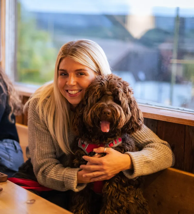 Woman smiling and cuddling a fluffy brown dog at a wooden table by the window, with another woman and food and drinks nearby