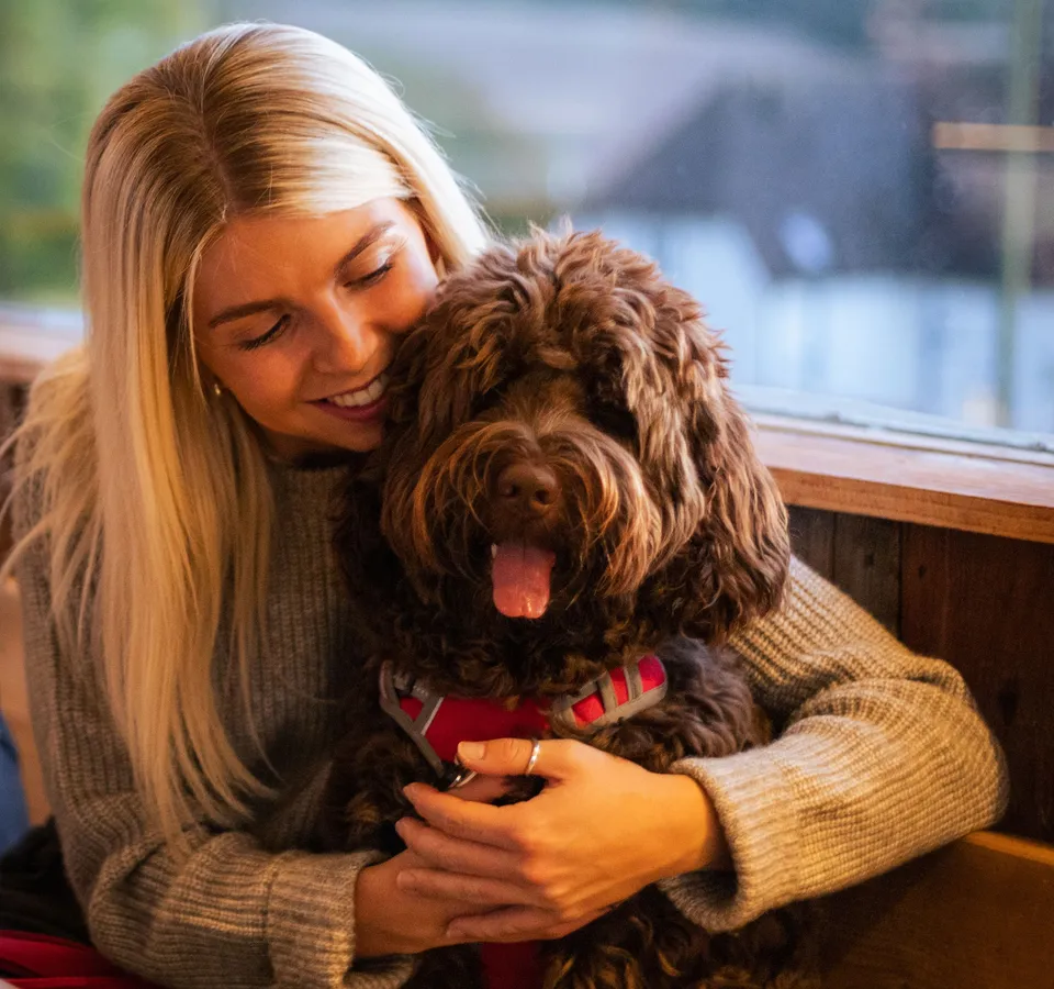 Woman cuddling a fluffy brown dog wearing a red harness by a window
