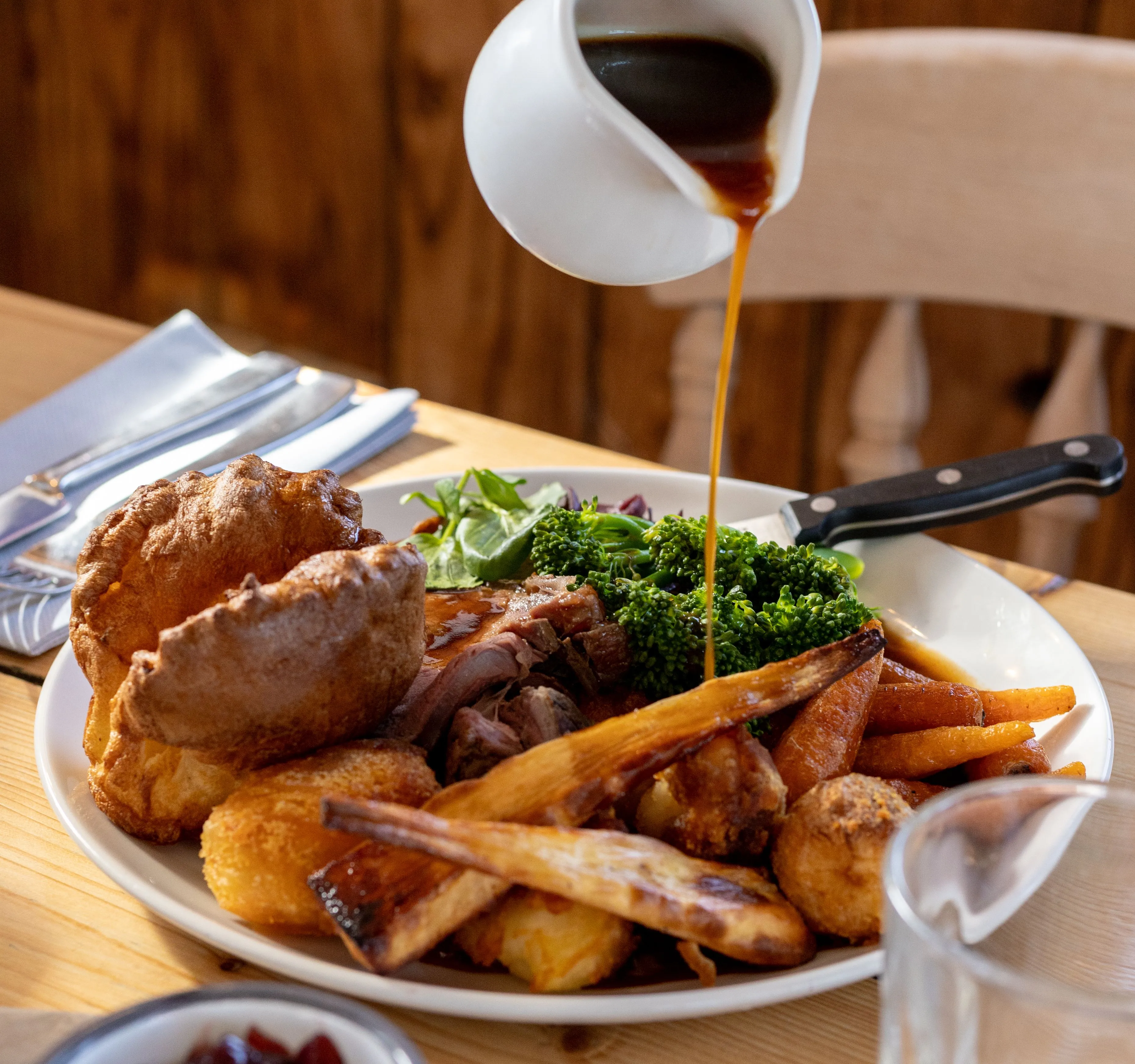 Gravy being poured over a Sunday roast with Yorkshire pudding, roast potatoes and vegetables on a wooden table