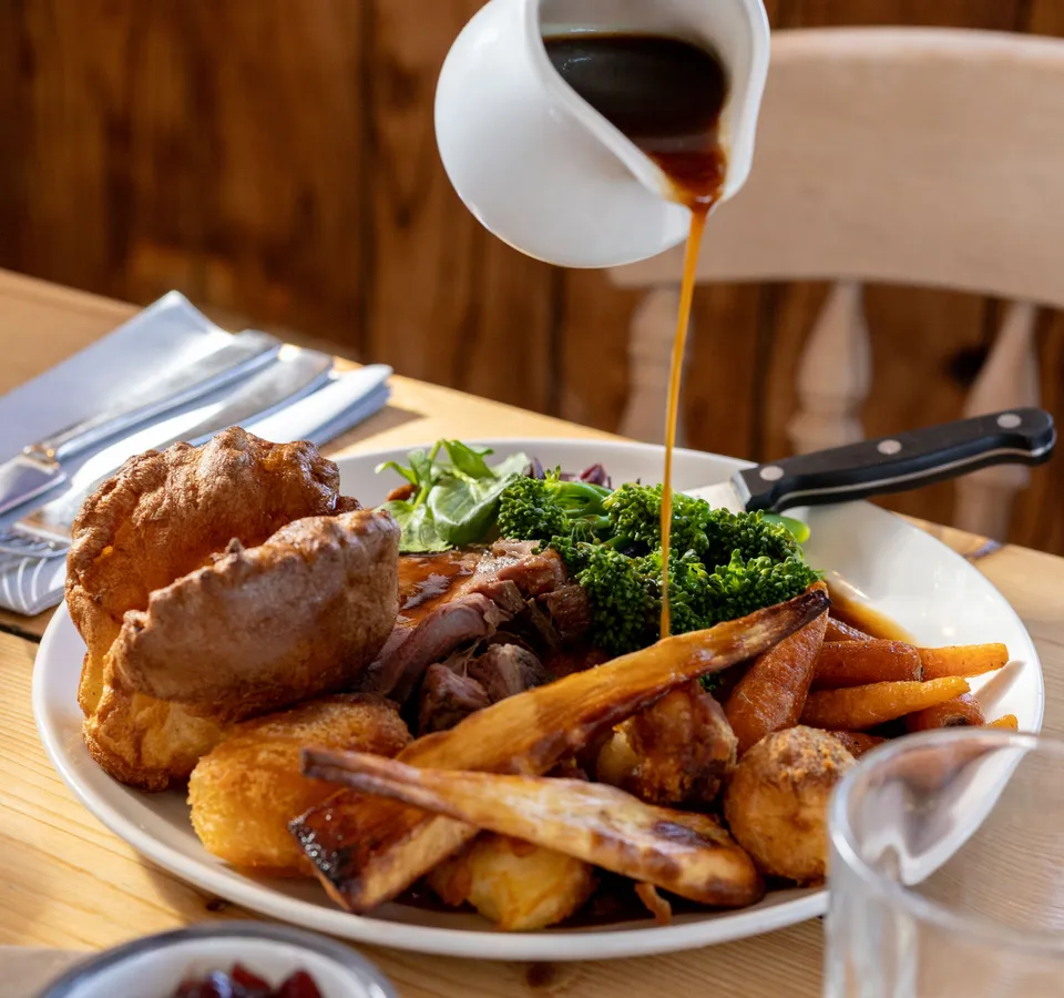 Gravy being poured over a Sunday roast with Yorkshire pudding, roast potatoes and vegetables on a wooden table