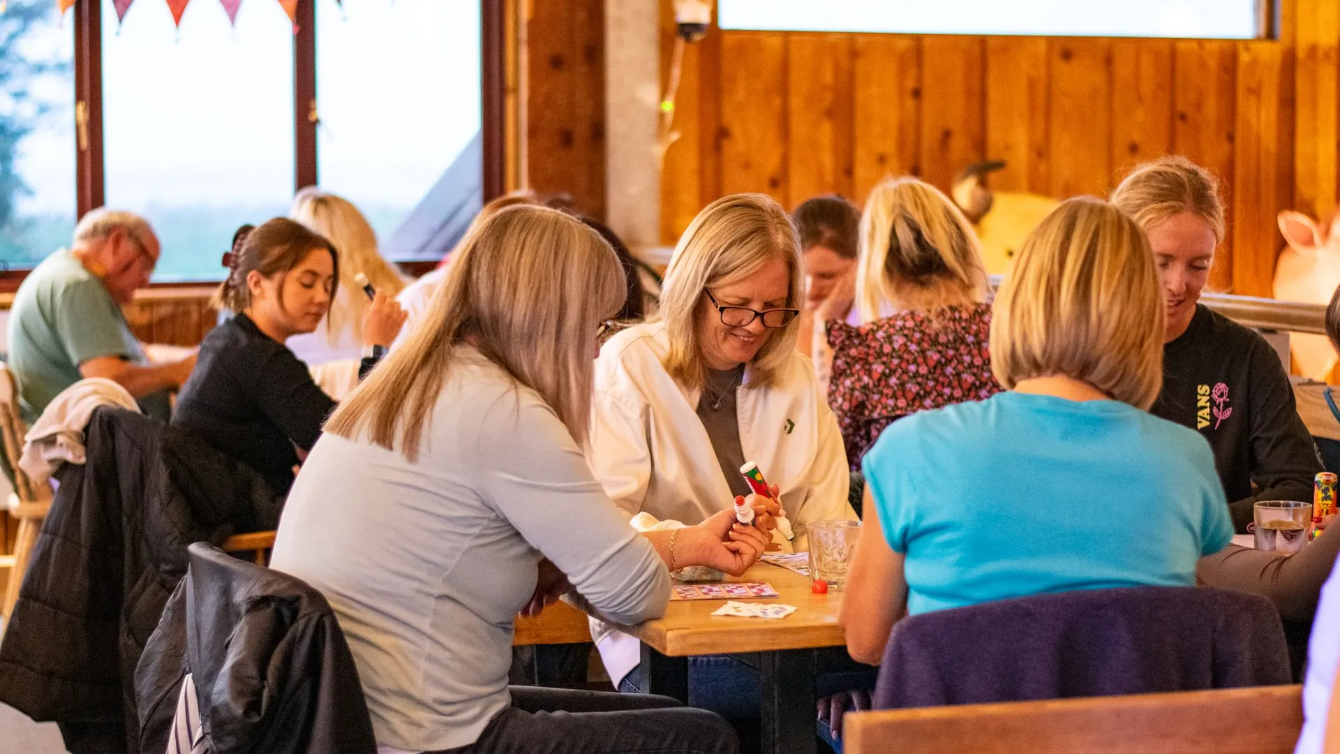 People playing bingo at tables in a warm, wooden hall with bunting overhead