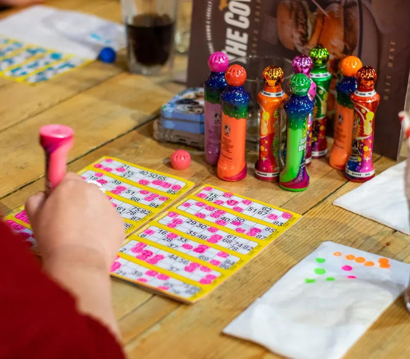 Hands marking bingo cards with a pink dabber on a wooden table, with colourful bingo dabbers and drinks nearby.