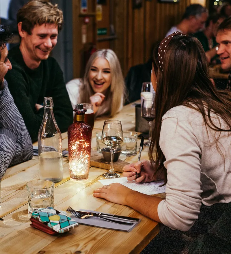 Friends laughing around a wooden table during a pub quiz night, with drinks and a woman writing answers on a sheet.