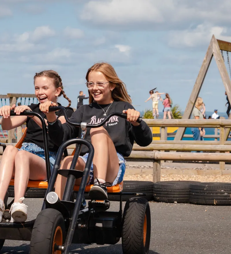 Two girls laughing while riding a pedal go-kart at an outdoor play area