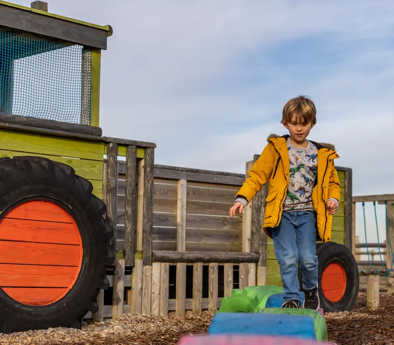 Child balancing on colourful tyre steps at an outdoor farm park play area beside a wooden tractor structure.