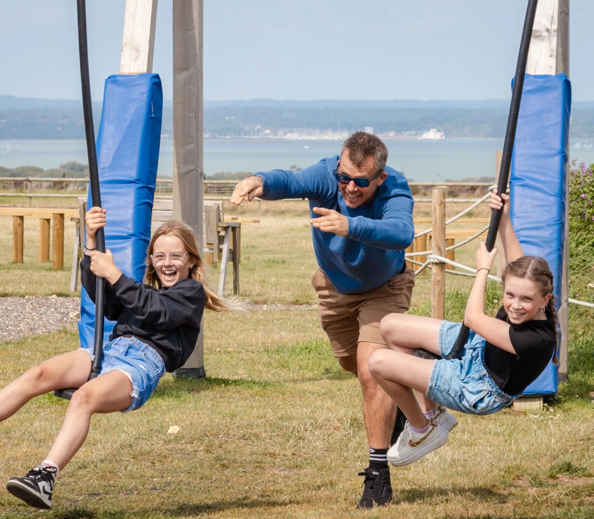 Two girls on a farm park zip line as an adult runs behind them, with coastal views in the background.