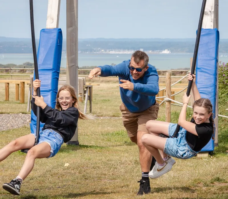 Two girls on a farm park zip line as an adult runs behind them, with coastal views in the background.