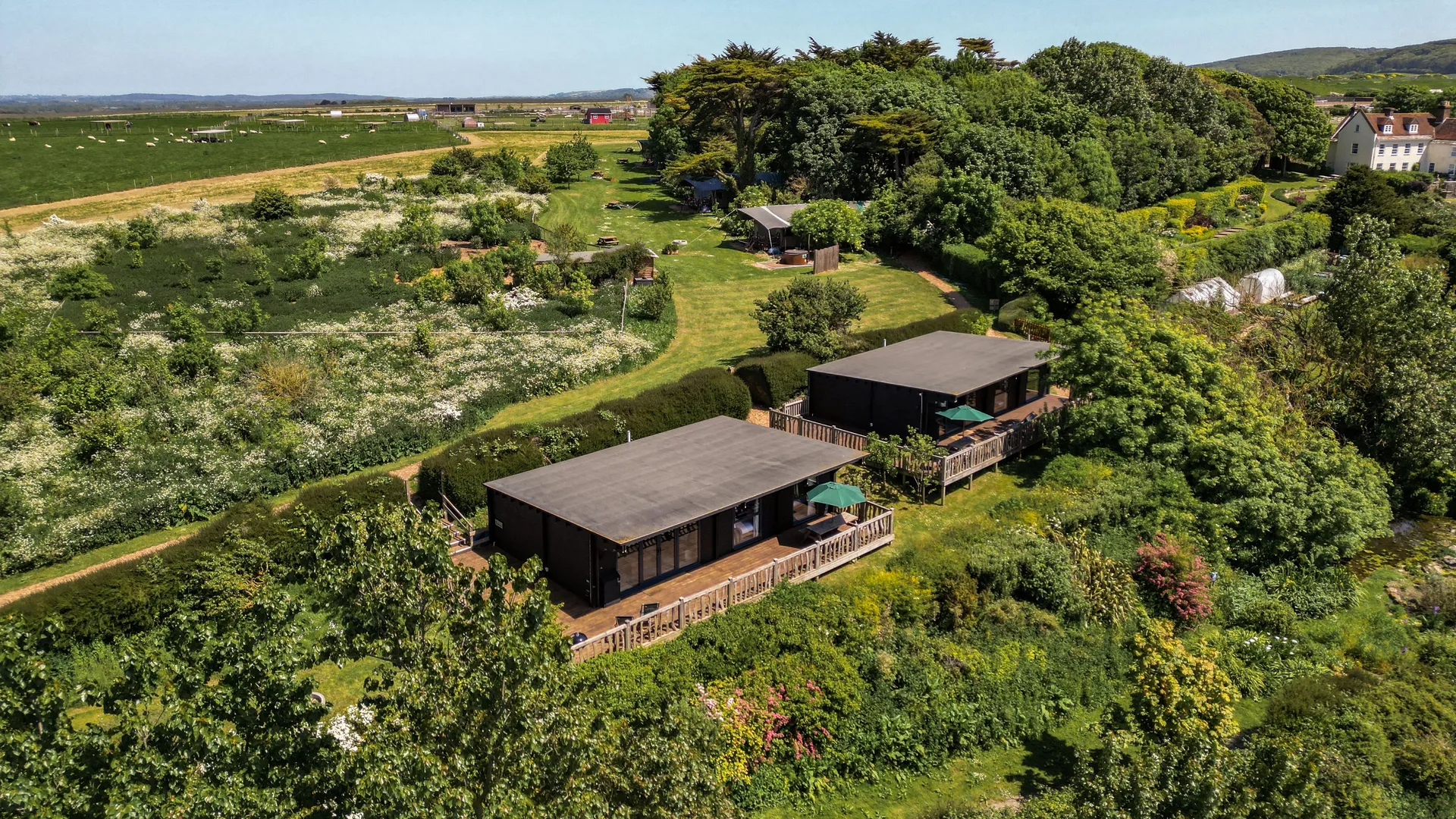 Aerial view of two modern wood cabins with decks set among lush gardens and fields under a clear blue sky