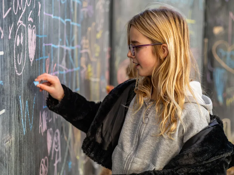 Young girl drawing with blue chalk on a large outdoor chalkboard.