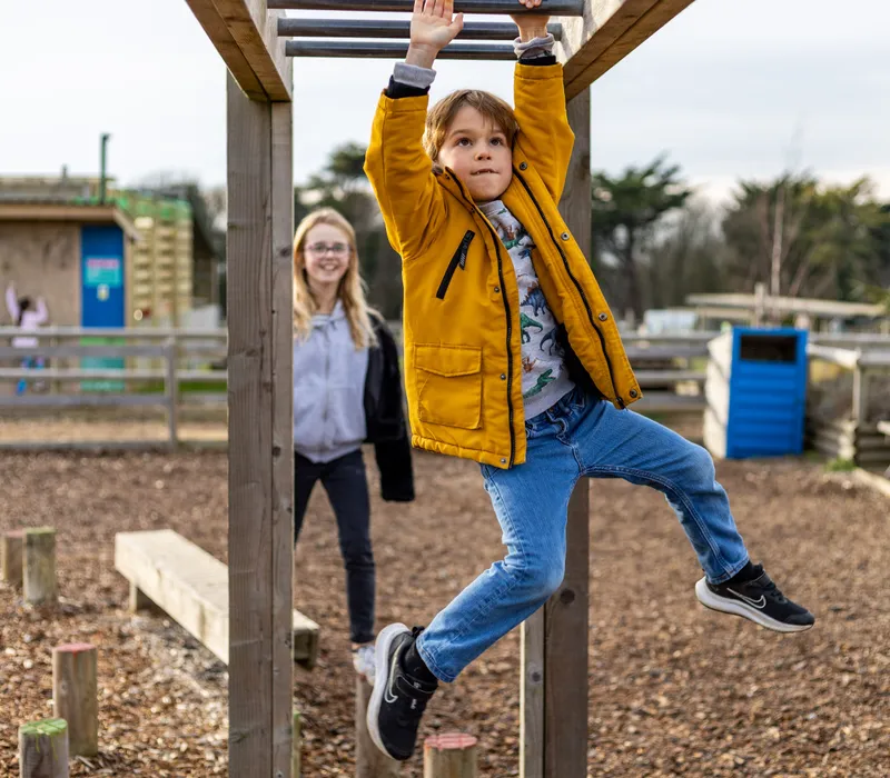 Child in a yellow coat hanging from monkey bars at an outdoor farm park playground